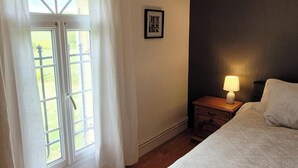 Un dormitorio con una cama, una mesita de noche con lámpara, una ventana con cortinas y un cuadro en la pared.