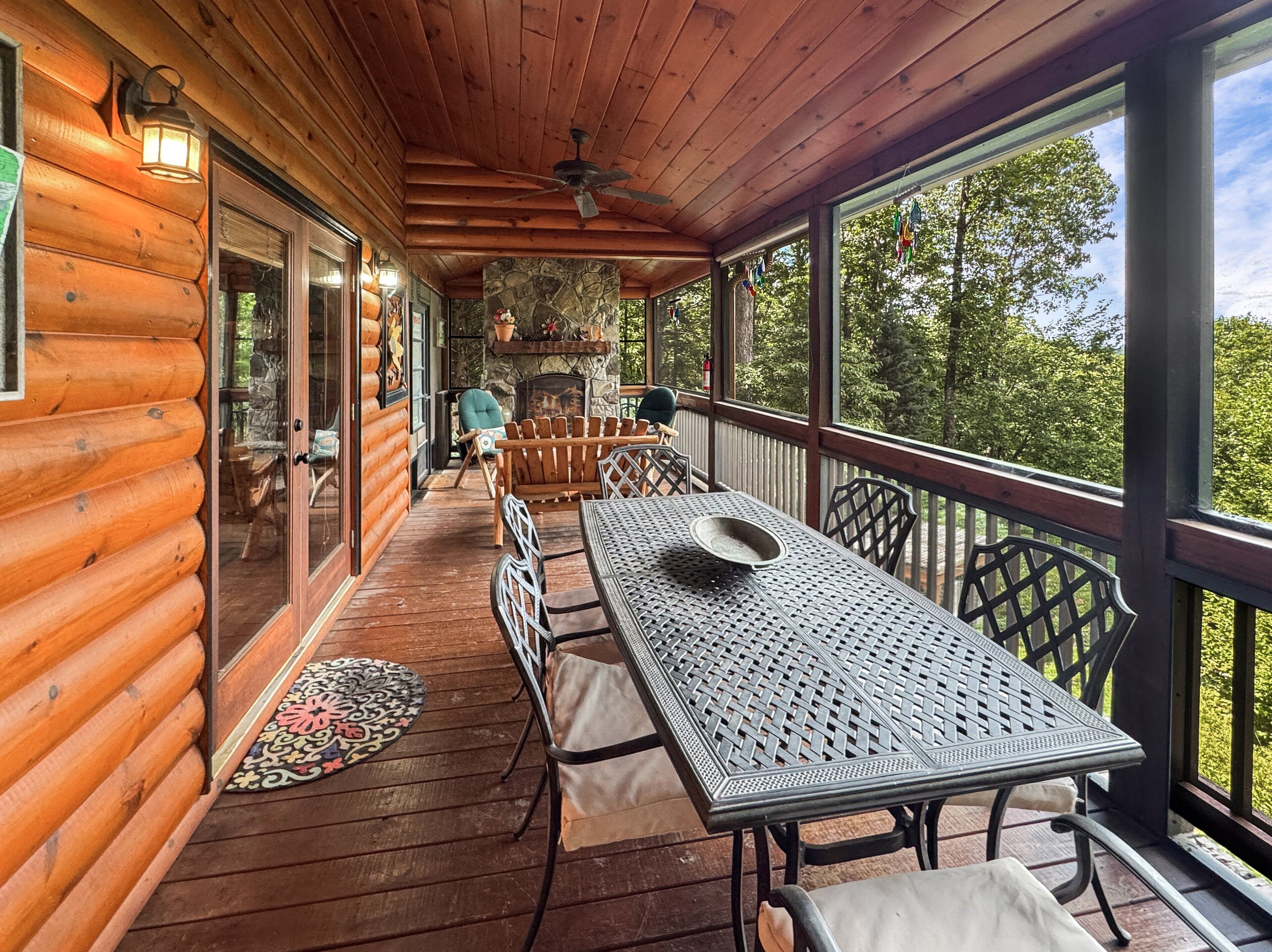 A wooden deck with a table and chairs, a stone fireplace, and a view of the surrounding forest.