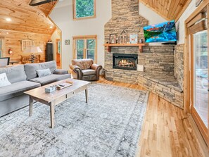 A cozy living room with a stone fireplace, wooden walls, a gray sofa, and a television mounted above the fireplace. - Mountain Melody - Mountain View | Pet Friendly | Hot Tub | Game Room (Morganton)