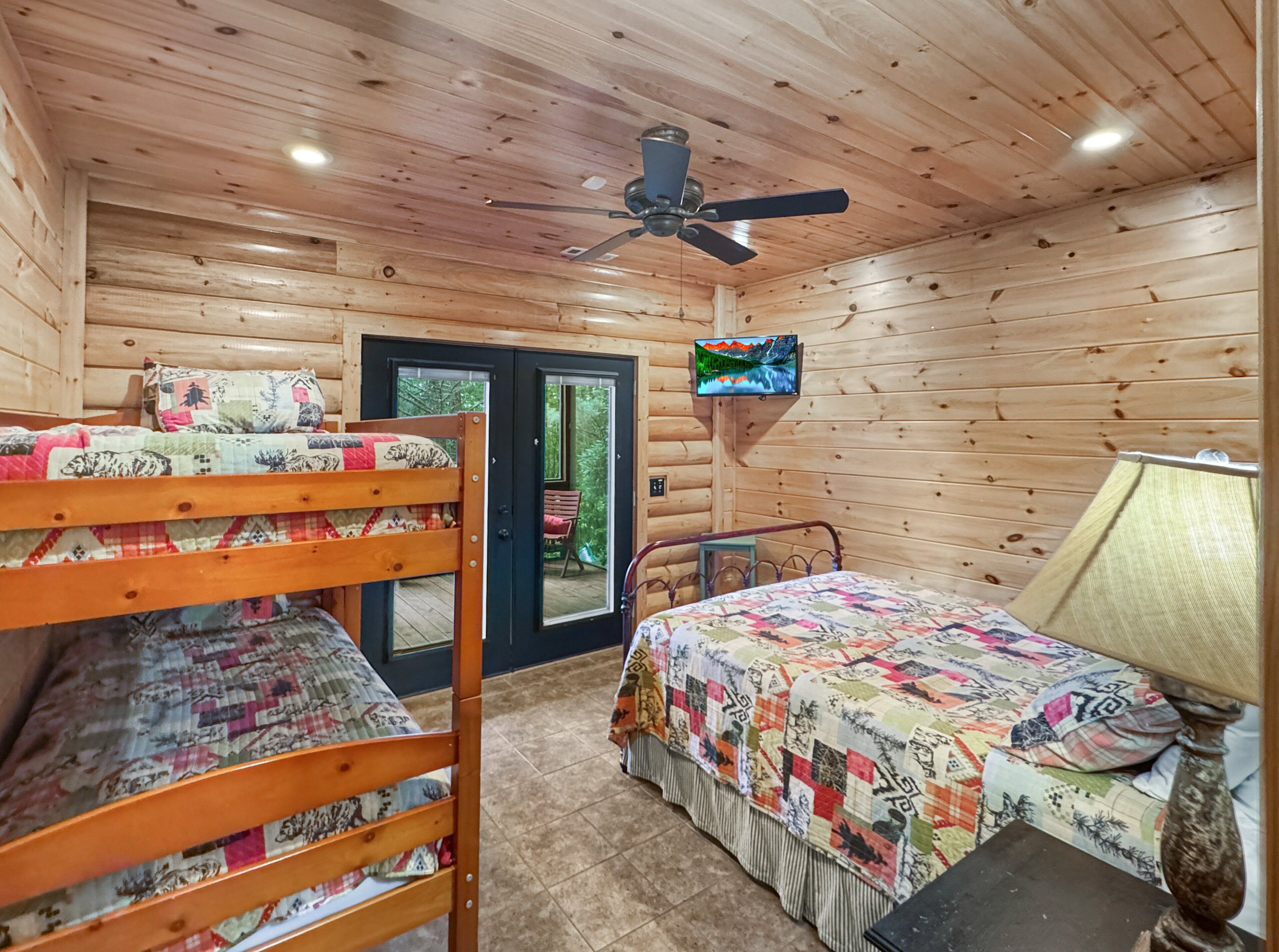 A cabin-style room with a bunk bed, a single bed, a ceiling fan, a television, and a door leading to an outdoor area.