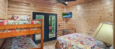 A cabin-style room with a bunk bed, a single bed, a ceiling fan, a television, and a door leading to an outdoor area.