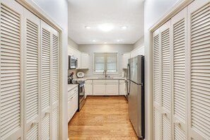 Ein schmaler Flur, am Ende befindet sich eine Küchenzeile mit weißen Schränken, einem Kühlschrank und einem Fenster mit Jalousien.