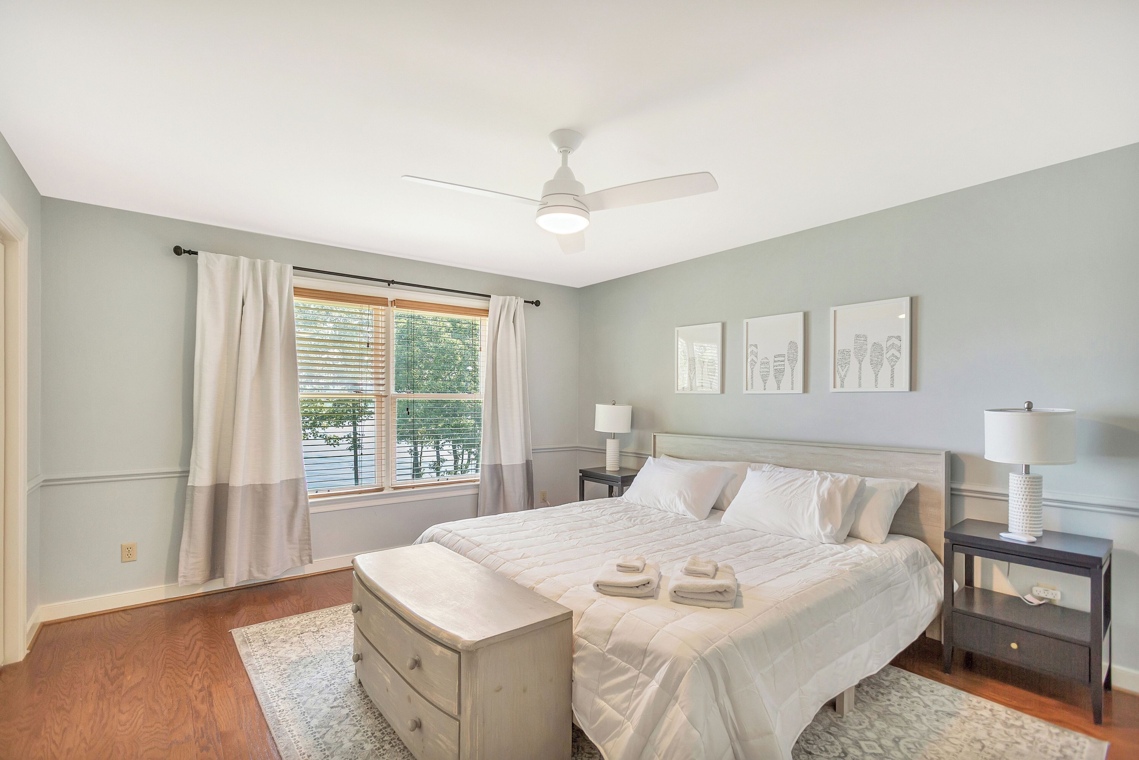 A bedroom with a bed, bedside tables, a ceiling fan, a window with curtains, and wall art.