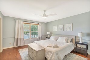 Une chambre à coucher comprenant un lit, des tables de chevet, un ventilateur de plafond, une fenêtre avec des rideaux et des œuvres d’art murales.
