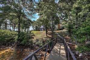 A wooden bridge leading to a house with a lawn and trees. - Eagles Point Waterfront Oasis (Seneca)