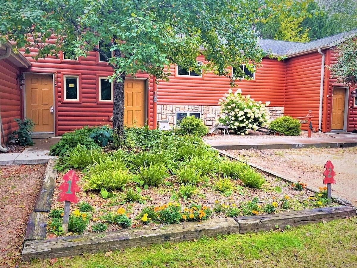 Una cabaña de madera roja con un jardín delantero, una cerca de madera y un buzón.