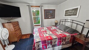 Quarto com cama, cômoda, televisão, ventilador, janela com cortinas e uma frase emoldurada na parede.