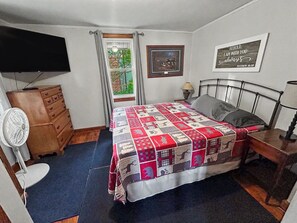 Ein Schlafzimmer mit Bett, Schminktisch, Fernseher, Ventilator, einem Fenster mit Vorhängen und einem gerahmten Zitat an der Wand.