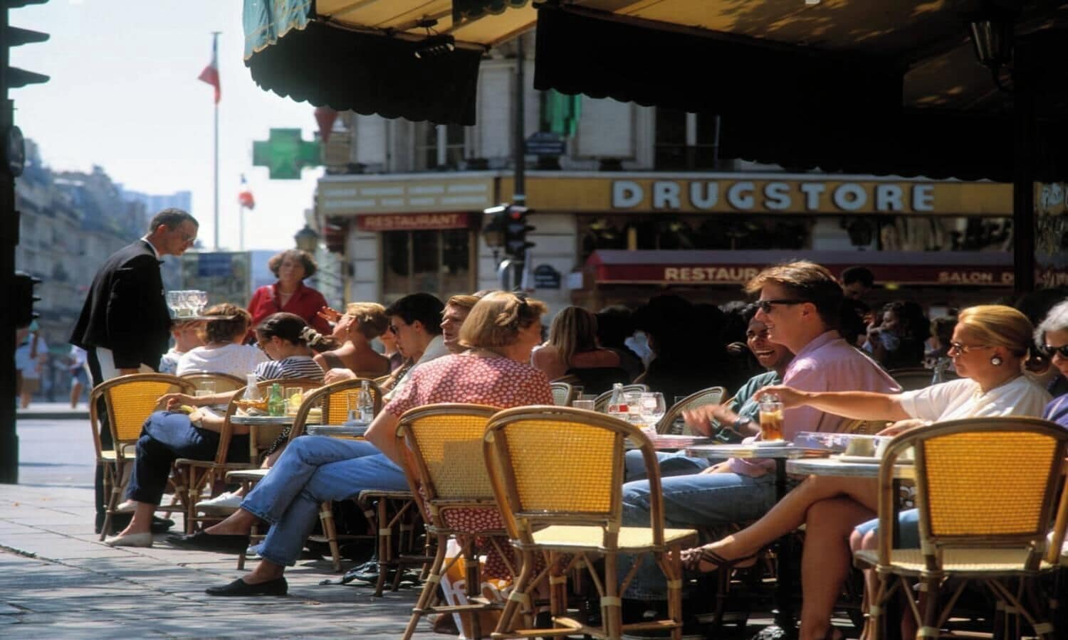 Un café animé, avec une terrasse, des chaises et des tables en osier, une pharmacie et un restaurant en arrière-plan.