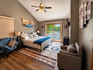 Ein Schlafzimmer mit einem Bett, einem blauen Sofa, einem grauen Sessel, einem Deckenventilator und Blick auf einen See durch eine Schiebetür.