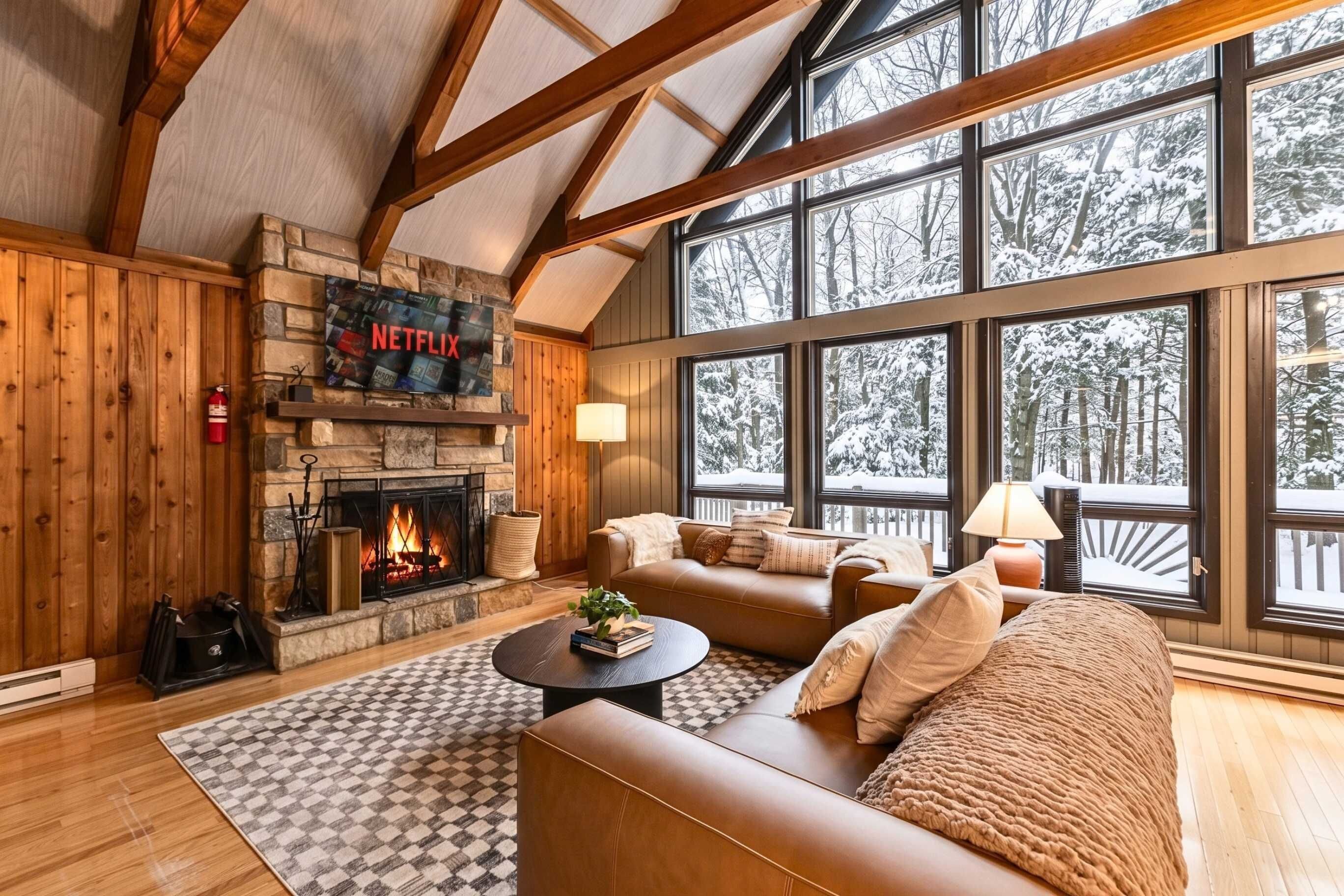 A cozy living room with a fireplace, leather sofas, a coffee table, and large windows with a snowy view.