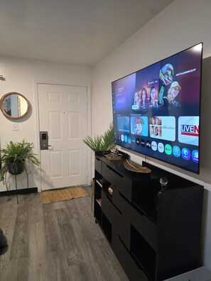Ein modernes Wohnzimmer mit Flachbildfernseher an der Wand, einer schwarzen Unterhaltungsanlage darunter, einem runden Spiegel und einem Blumentopf.