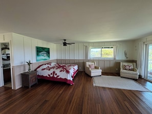 A bedroom with a bed, two chairs, a ceiling fan, a window, and a painting on the wall.