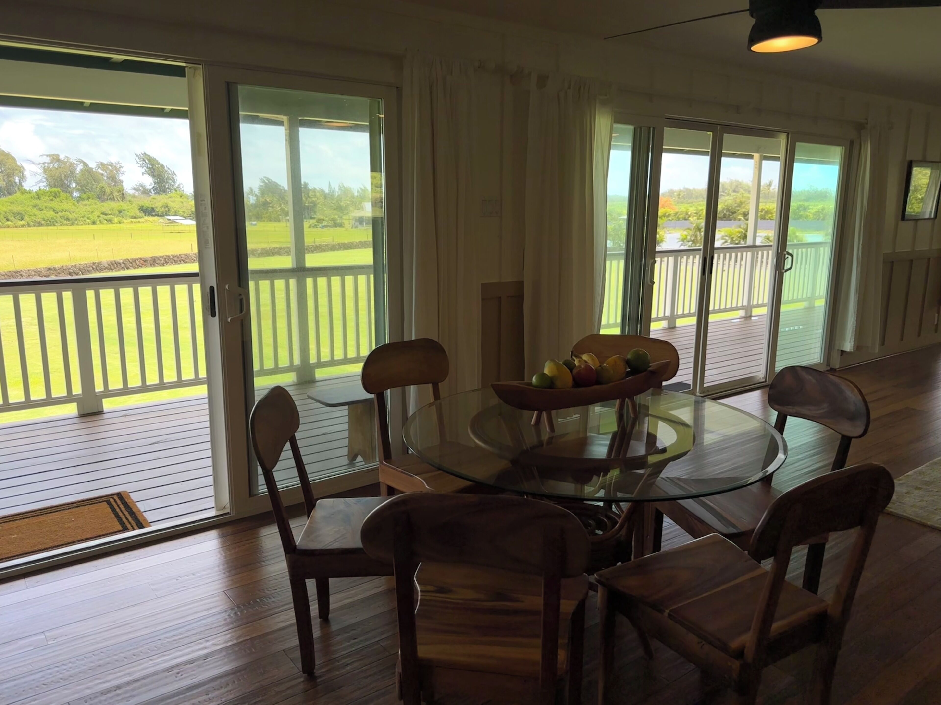 Un coin repas avec une table et des chaises en verre, un saladier rempli de fruits et une vue sur un paysage verdoyant, visible à travers de grandes portes coulissantes.