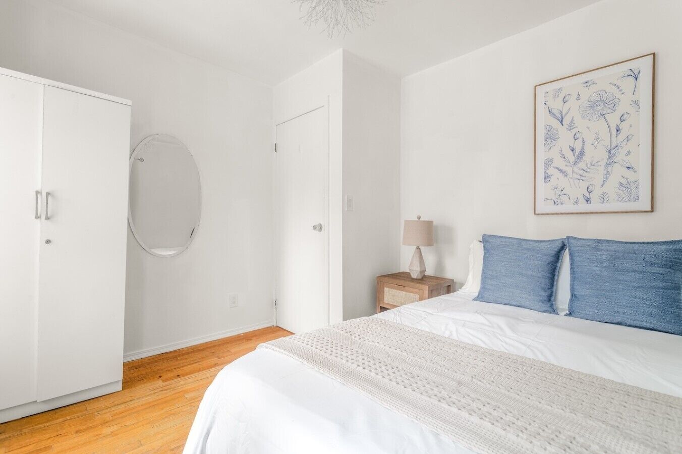 A bedroom with a bed, a nightstand, a lamp, a framed floral artwork, and a wardrobe.