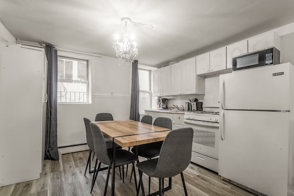 A compact kitchen with a dining table, chairs, a chandelier, and a refrigerator.