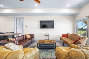 A spacious living room with a brown leather sofa, a patterned armchair, a wooden coffee table, and a large window with a view of greenery.