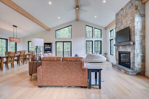 A spacious living room with a stone fireplace, large windows, and a dining area with wooden furniture.