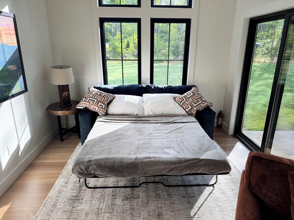 A bedroom with a bed, a nightstand, a lamp, and a view of the outdoors through large windows.