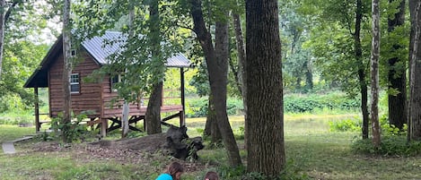 A rustic wooden cabin with a deck, surrounded by a forested area with a pond.
