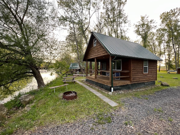 A wooden cabin with a porch, a picnic table, and a small shed by a body of water.