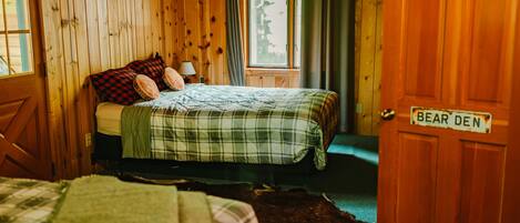 Interior de una cabaña con cama, una puerta de madera con un letrero y una ventana con cortinas.