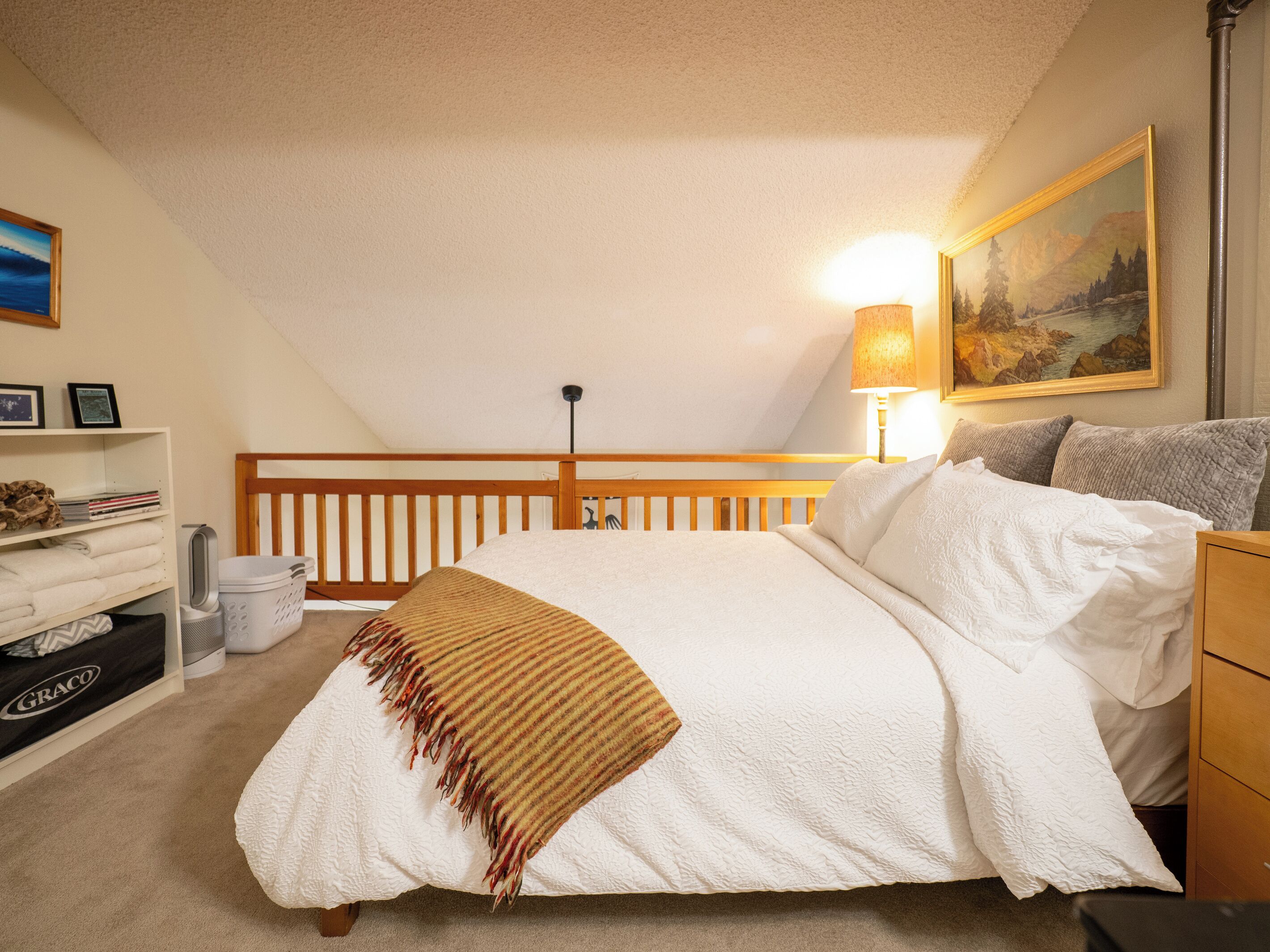 A bedroom with a sloped ceiling, a bed with white bedding, a wooden headboard, a nightstand, a lamp, and a painting on the wall.