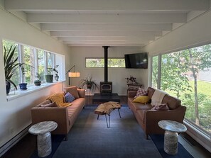 Un salón con dos sofás, una mesa de centro y una chimenea. Hay ventanas grandes a ambos lados, y un televisor montado en la pared.