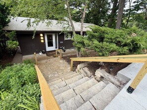 A residential property with a stone staircase leading to a wooden deck, surrounded by lush greenery and a dark-colored house. - 4-bedroom house in Iowa City-walking distance to Kinnick, Carver and UIHC (Iowa City)