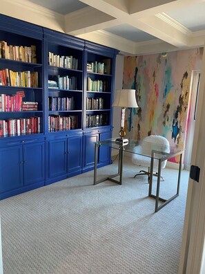 Ein Zimmer mit einem blauen Bücherregal, das mit Büchern gefüllt ist, einem Glastisch mit Lampe und einem weißen Stuhl.