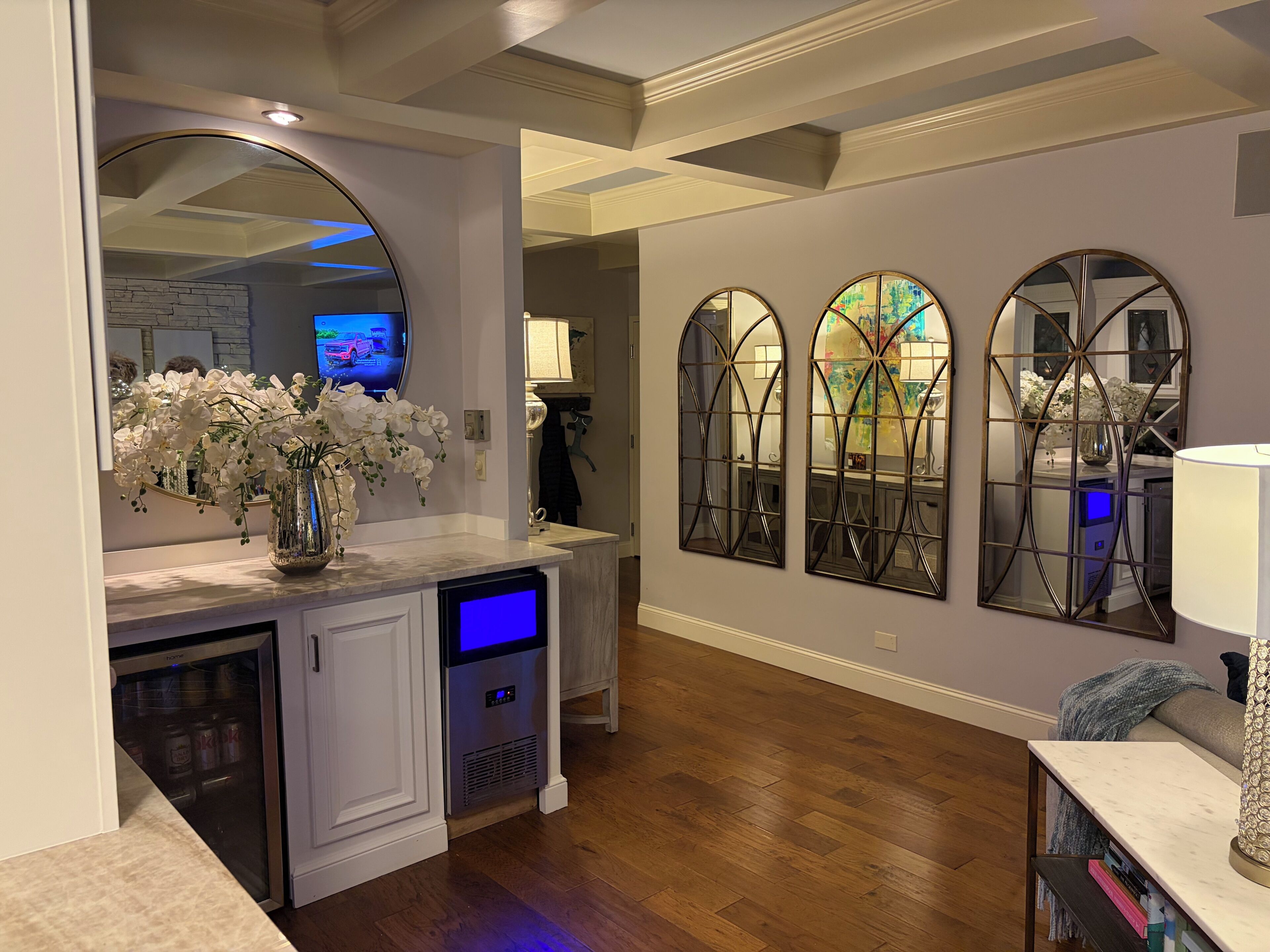 A modern kitchen with a built-in refrigerator, a microwave, and a coffee maker. There are three wall-mounted mirrors and a vase of white flowers on the counter.