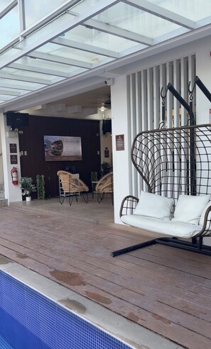 A covered outdoor seating area with wicker chairs and a swing chair, a poolside, and a building entrance with a sign.