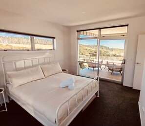 A bedroom with a large bed, a view of a deck with chairs, and a door to the right.