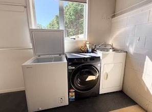 A compact laundry room with a front-loading washer and dryer, a small sink, and a window providing natural light. - Mānuka Retreat - Private Luxury, Panoramic Views (Cable Bay)