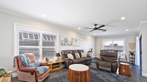 A living room with a sofa, armchair, and a coffee table. There are two windows with blinds, a ceiling fan, and artwork on the wall. - Serenity Place - Relax, Unwind & Explore at Serenity Place (Mannering Park)