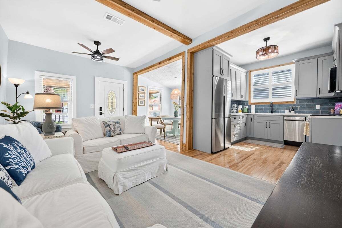 A modern kitchen with stainless steel appliances, a dining area with a white table and chairs, and a living room with a white sofa and a ceiling fan.