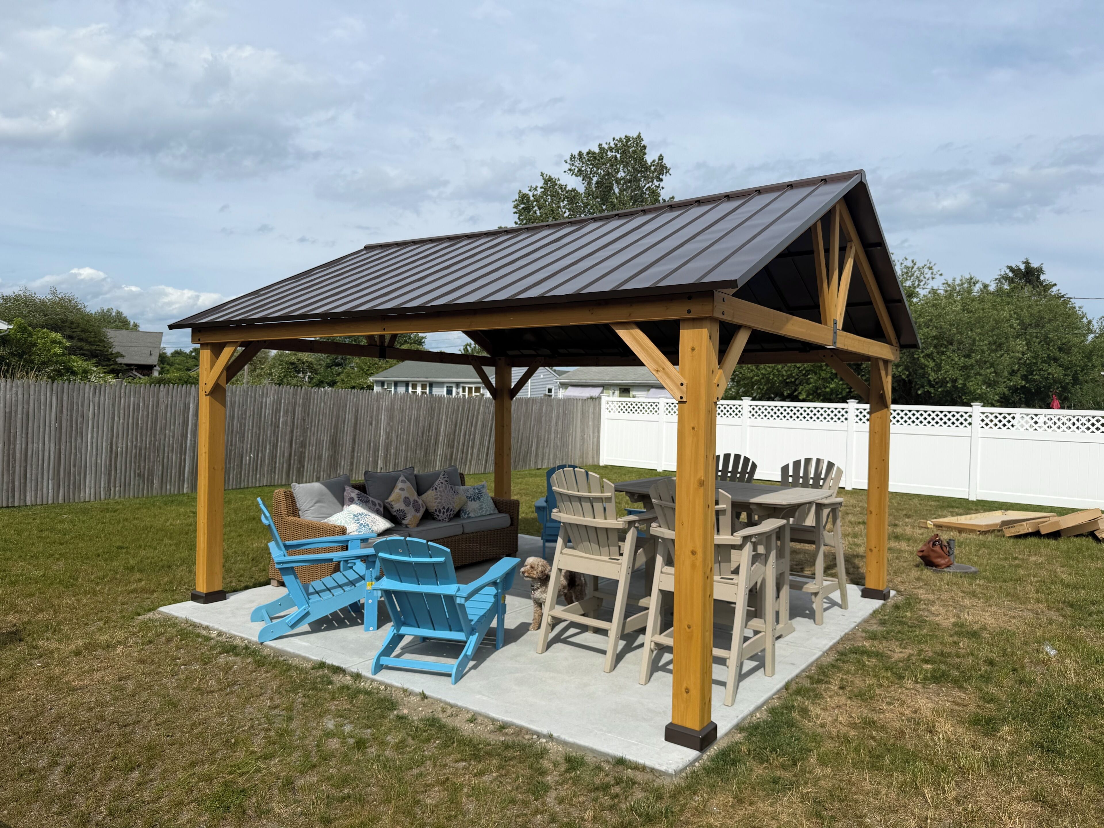 Un patio techado con estructura de madera, mobiliario exterior y un jardín cercado.