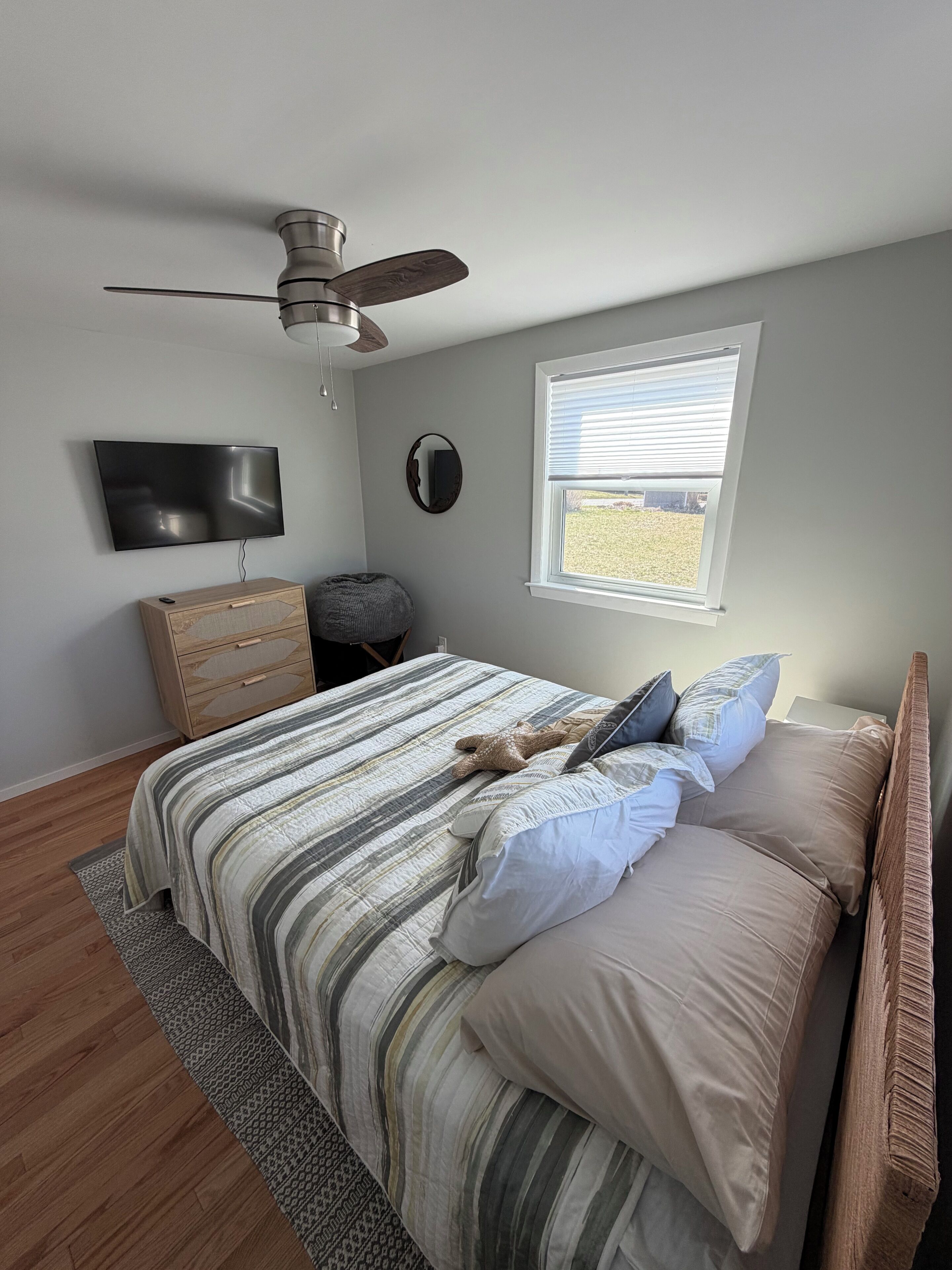 A bedroom with a bed, a ceiling fan, a TV, a window, and a bedside table.
