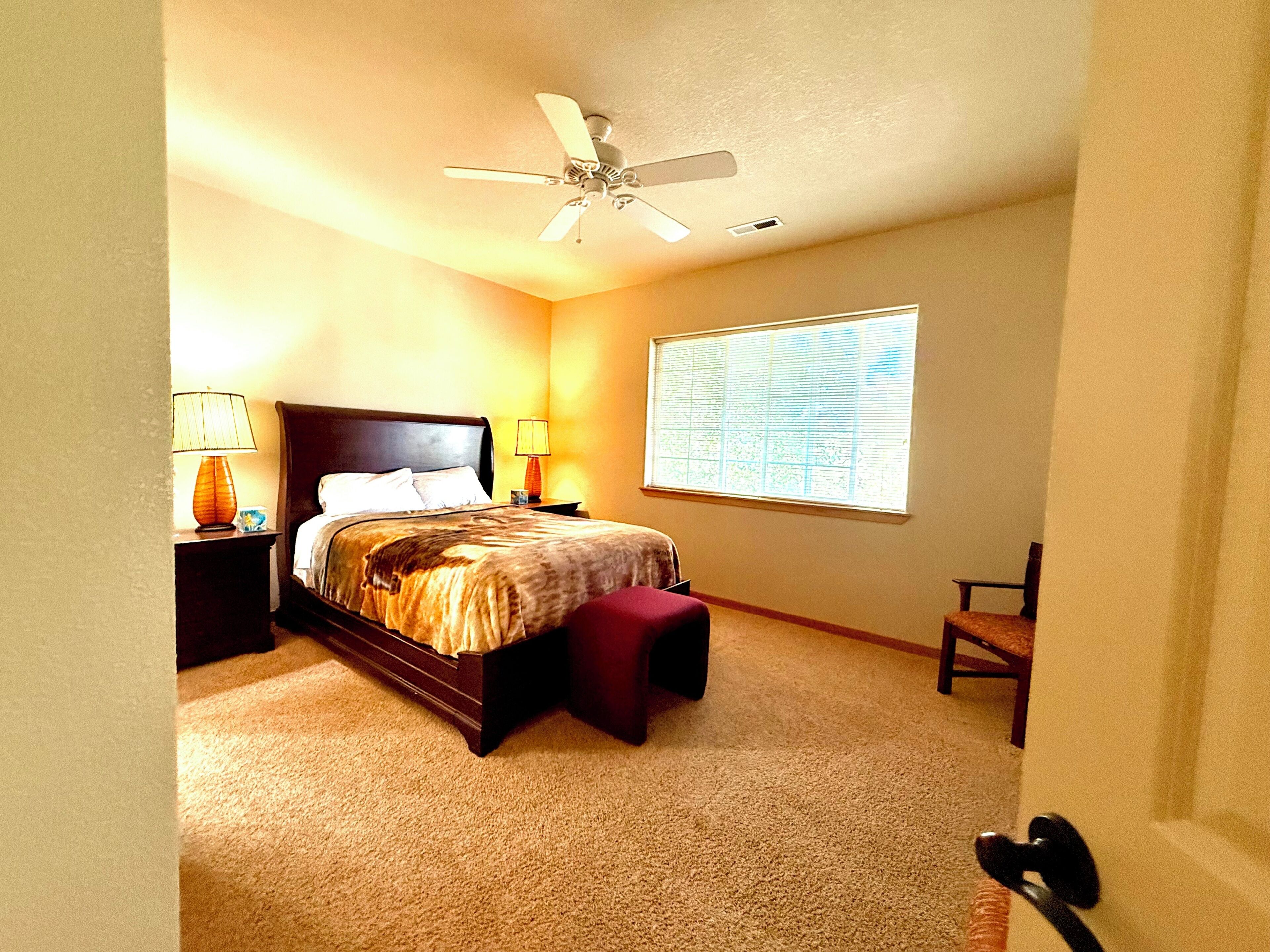 Un dormitorio con cama, mesita de noche, lámpara, silla, ventana y ventilador de techo.