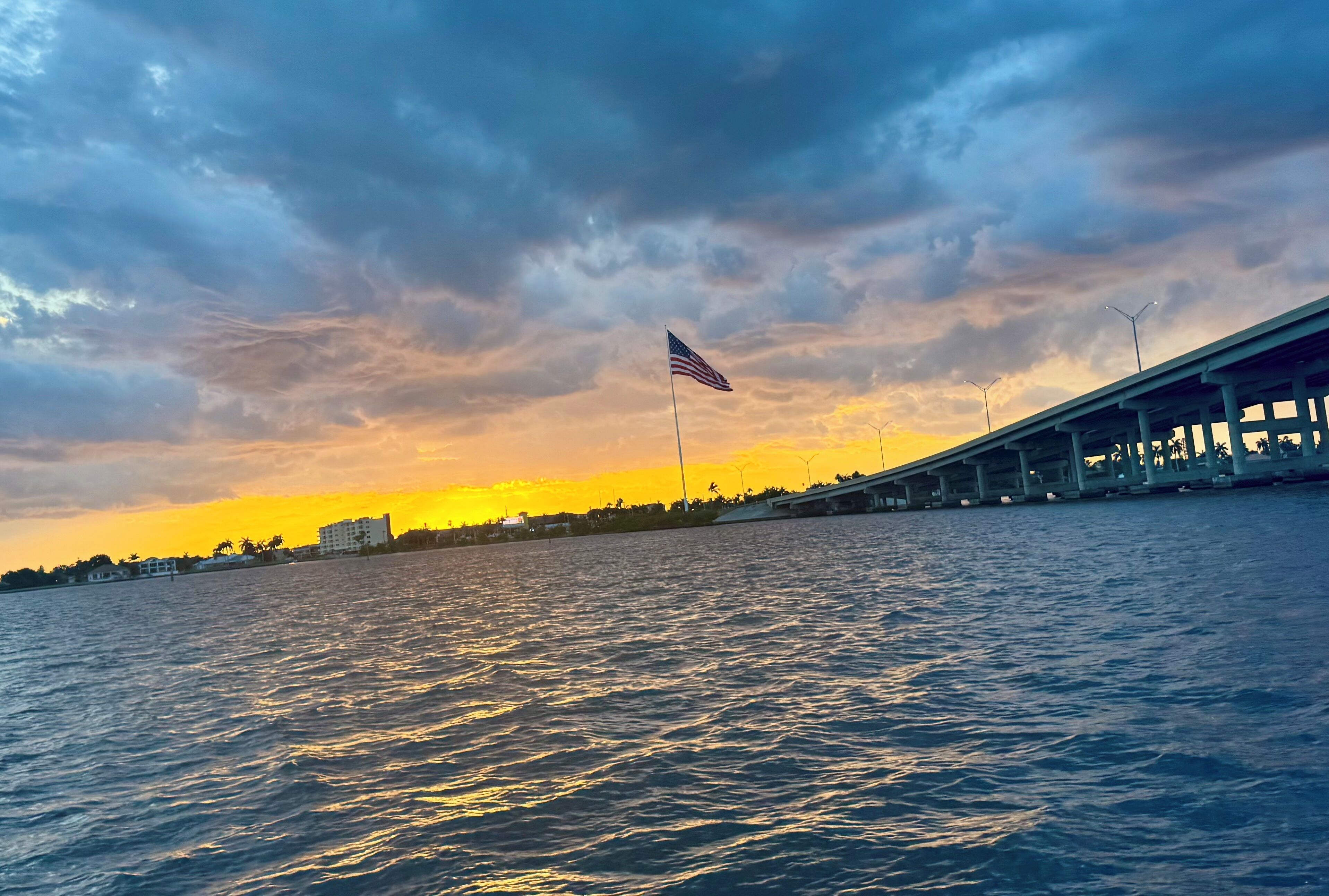A waterfront view with a bridge, an American flag, and a sunset.