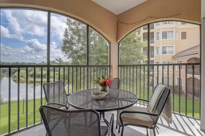 A covered outdoor patio with a round table and chairs, a vase with flowers, and a view of a lake and buildings. - Disney Doorstep Resort Vibes Stay. (Orlando)