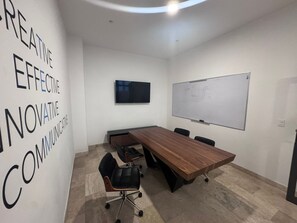 Ein Konferenzraum mit langem Holztisch, schwarzen Stühlen, einer Whiteboard-Tafel und einem Fernseher an der Wand.