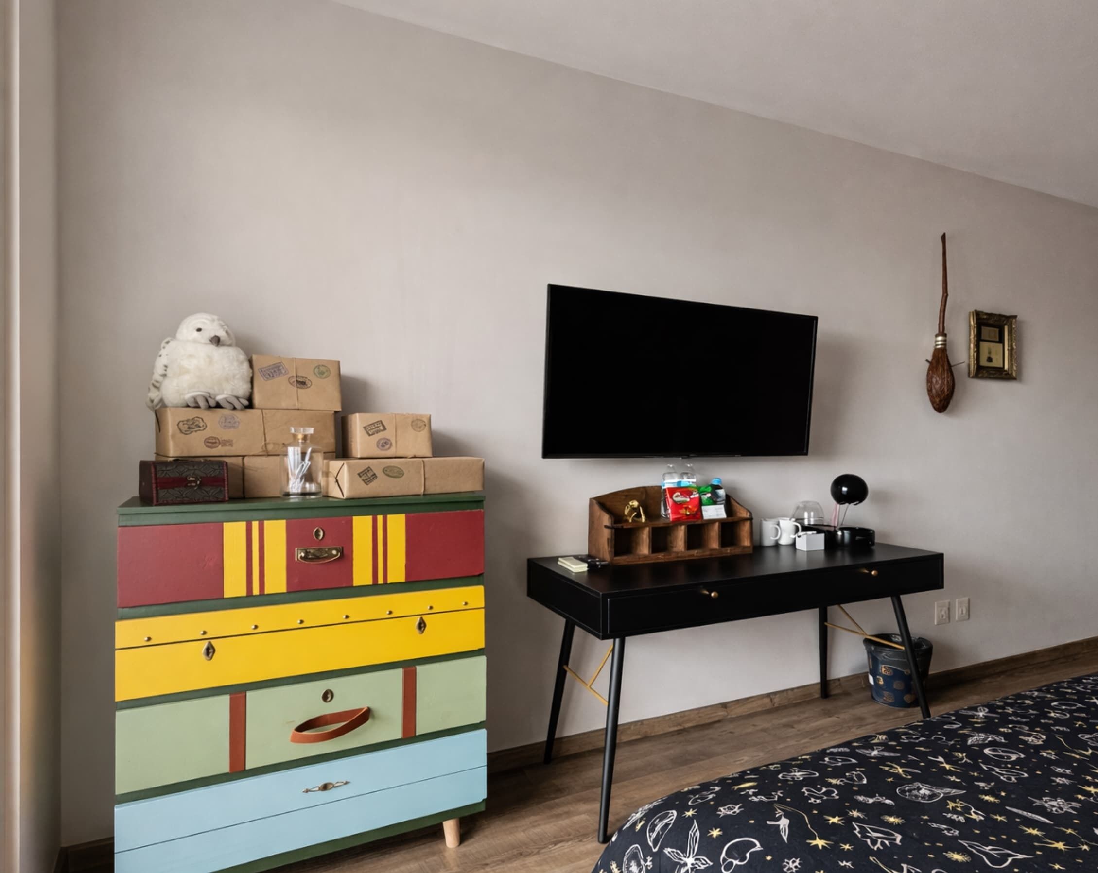 A room with a colorful dresser, a black desk with a flat-screen TV, and a bed with a patterned bedspread.