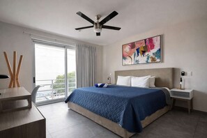 A hotel room with a bed, a desk, a chair, a ceiling fan, a painting, and a view through a window. - Stylish 1BR Condo w Rooftop Pool Near 5th Ave (Playa del Carmen)
