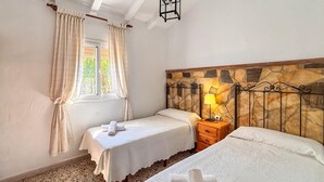A bedroom with two beds, a stone headboard, a window with curtains, and a nightstand with a lamp. - Amazing home in Frigiliana (Frigiliana)