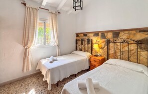 A bedroom with two beds, a stone headboard, a window with curtains, and a nightstand with a lamp. - Amazing home in Frigiliana (Frigiliana)