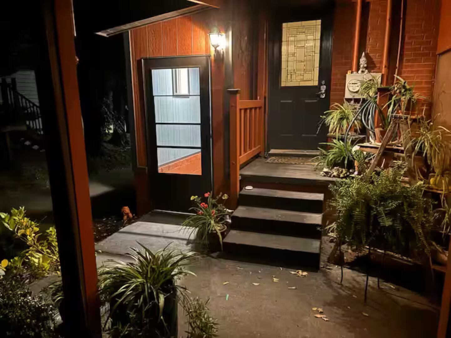 Entrada de una casa de madera con una puerta oscura, escalones que conducen a un porche y plantas en macetas.