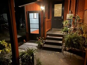 Entrada de una casa de madera con una puerta oscura, escalones que conducen a un porche y plantas en macetas.
