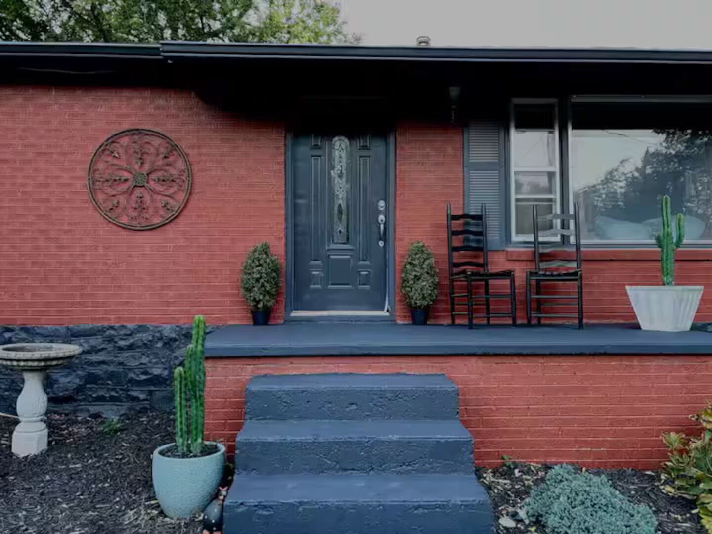 Uma casa de tijolos vermelhos com uma porta azul escura, dois vasos de plantas, uma mesa pequena e uma cadeira na parte externa.
