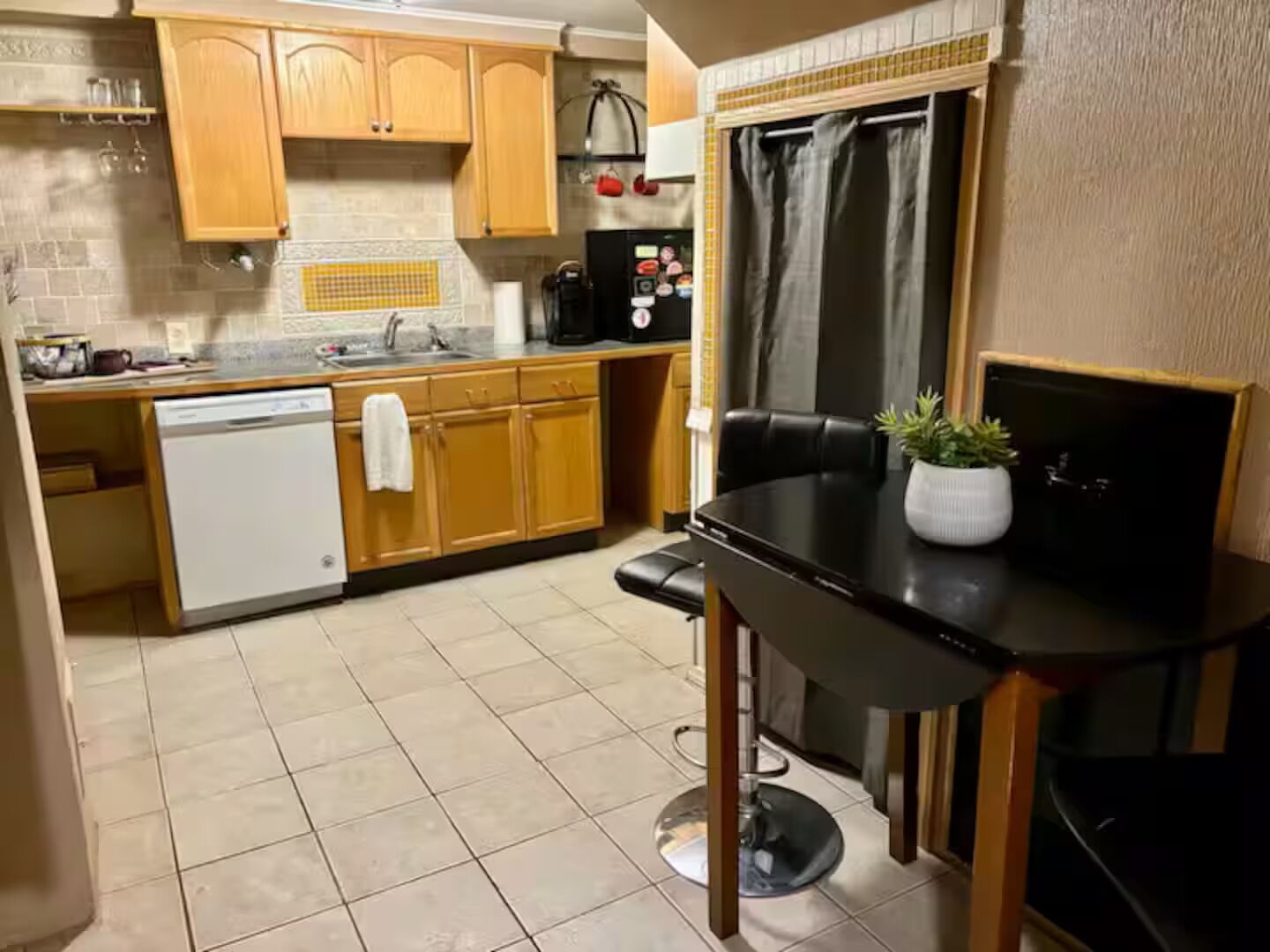 Una cocina compacta con armarios de madera, lavavajillas, cafetera y una pequeña mesa de comedor con planta en maceta.
